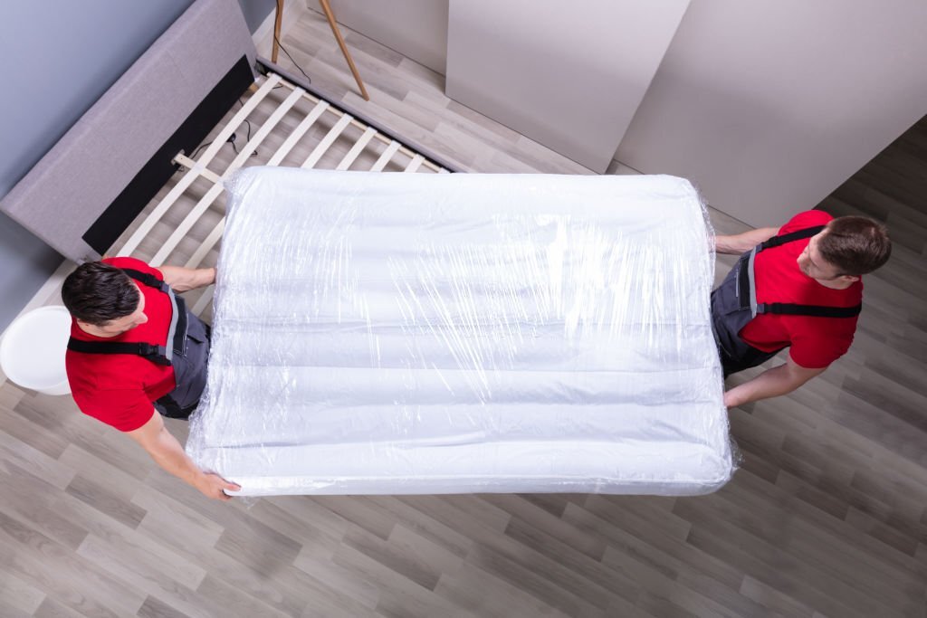 Two Male Professional Movers In Uniform Placing The Mattress Over The Bed In New House